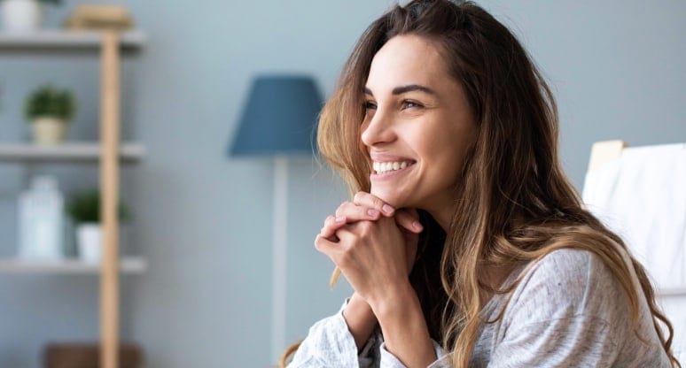 Woman with long brown hair smiling, her hands clasped under her chin
